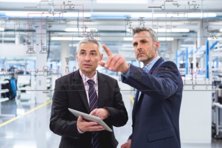 Zwei Geschäftsleute betrachten eine Grafik auf einer Glasscheibe in einer Fabrikhalle