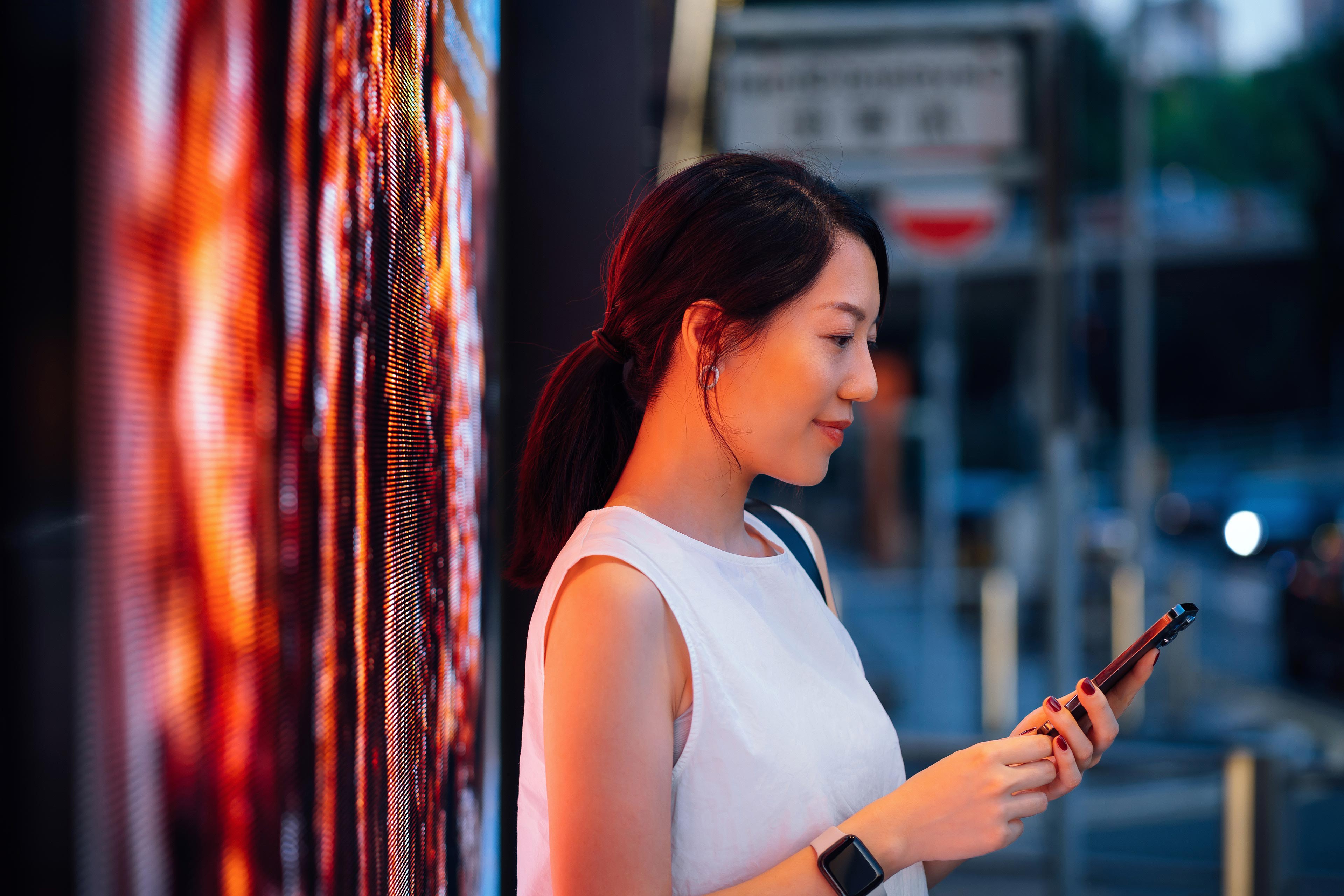 Confident young Asian businesswoman using smartphone in downtown city street in the evening, standing against illuminated LED digital display in the city. Lifestyle and technology
