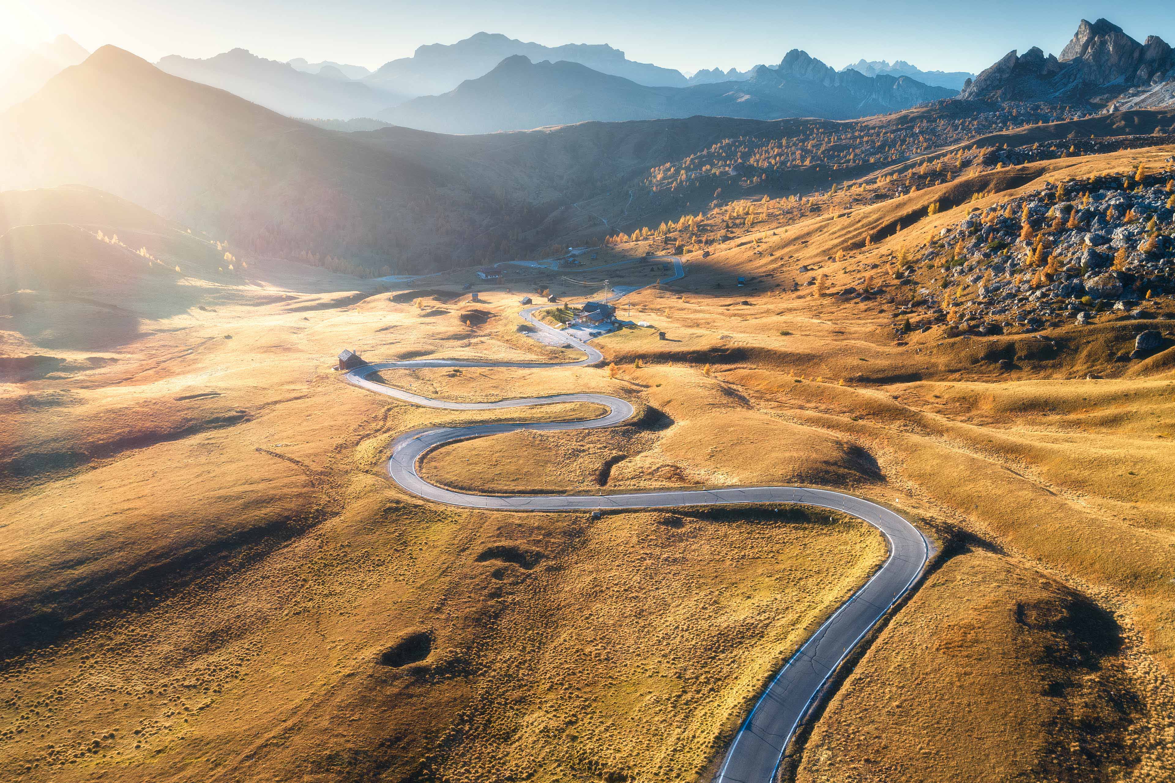 Winding road in mountain valley at sunset in autumn. Aerial view of asphalt road in Passo Giau. Dolomites, Italy. Top view of roadway, mountains, meadows with orange grass, blue sky and gold sunlight