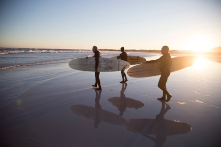 Three senior surfers, aged 55-70, walking along a sandy beach and preparing to go surfing