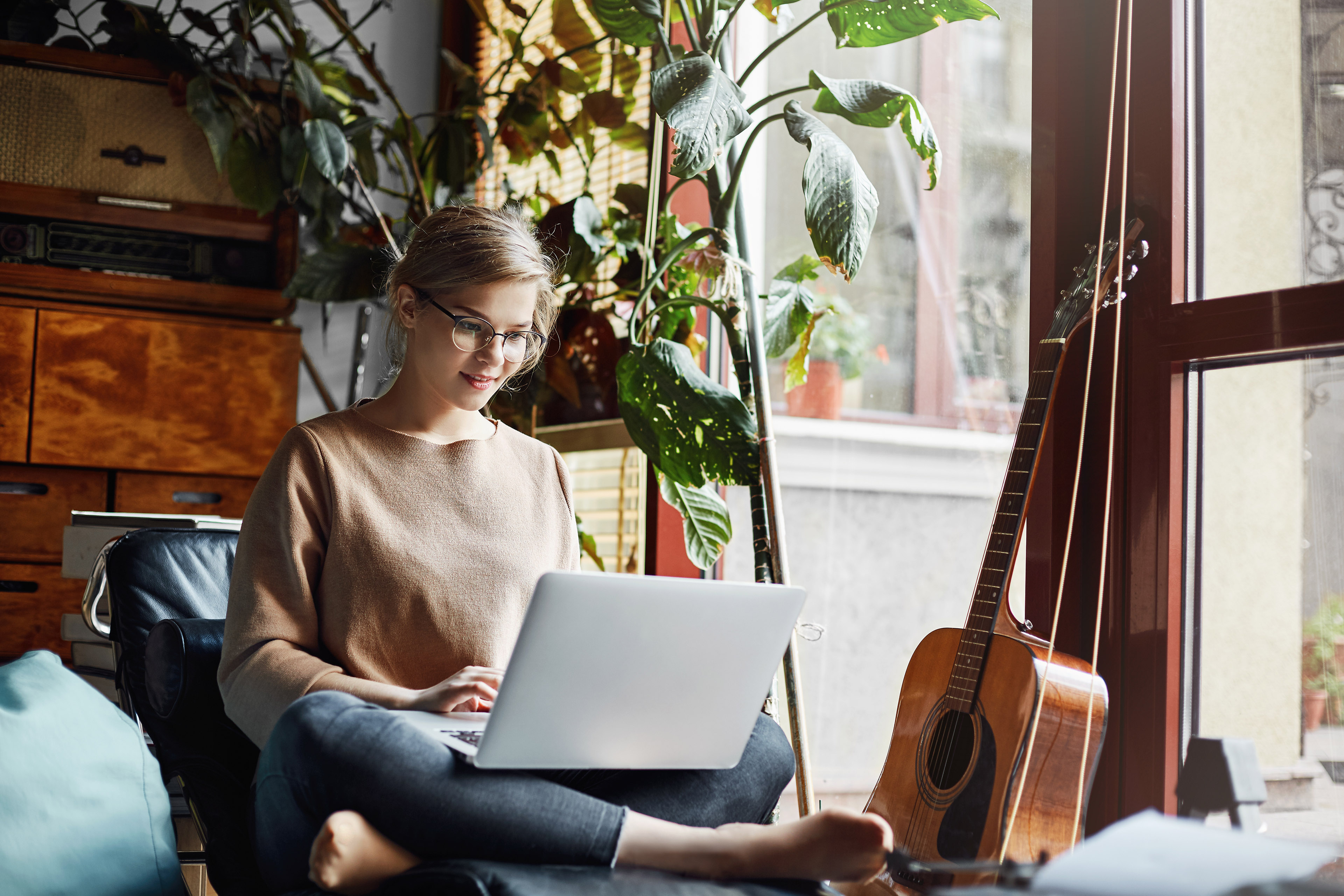 Girl sitting in cozy room near guitar and plant working on computer