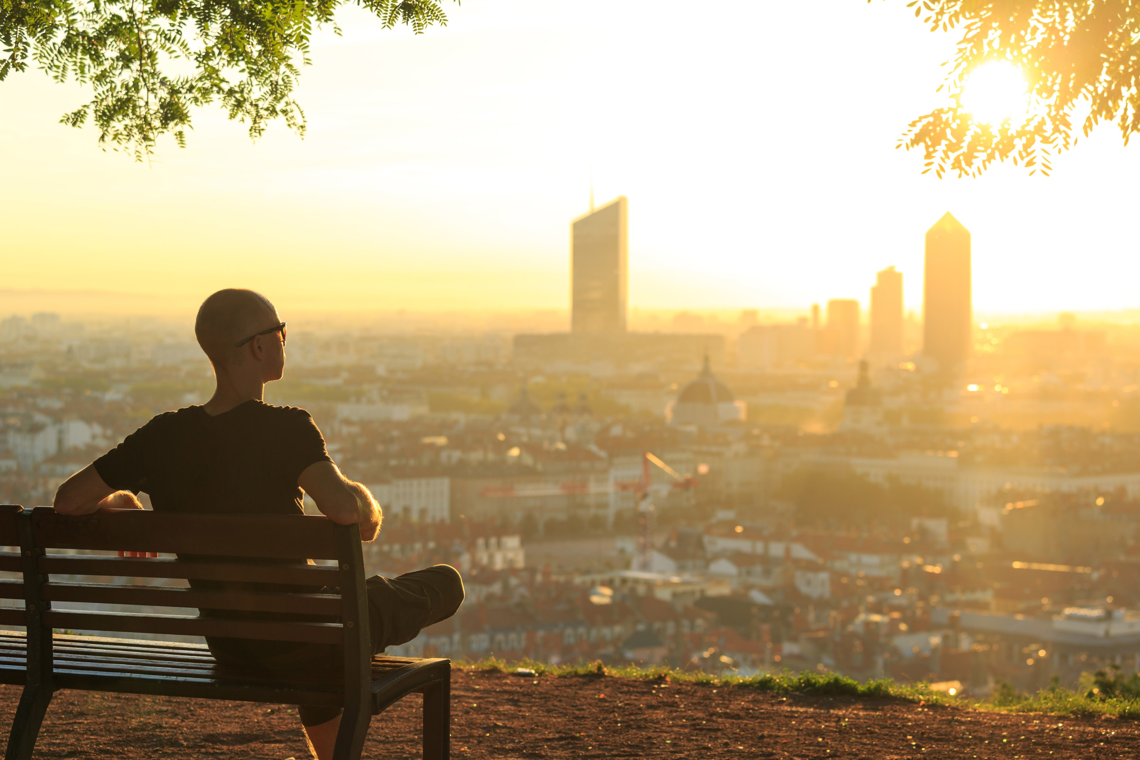 A man on a bench in a park, relaxing and enjoying the summer sunrise over a city. Lyon, France.