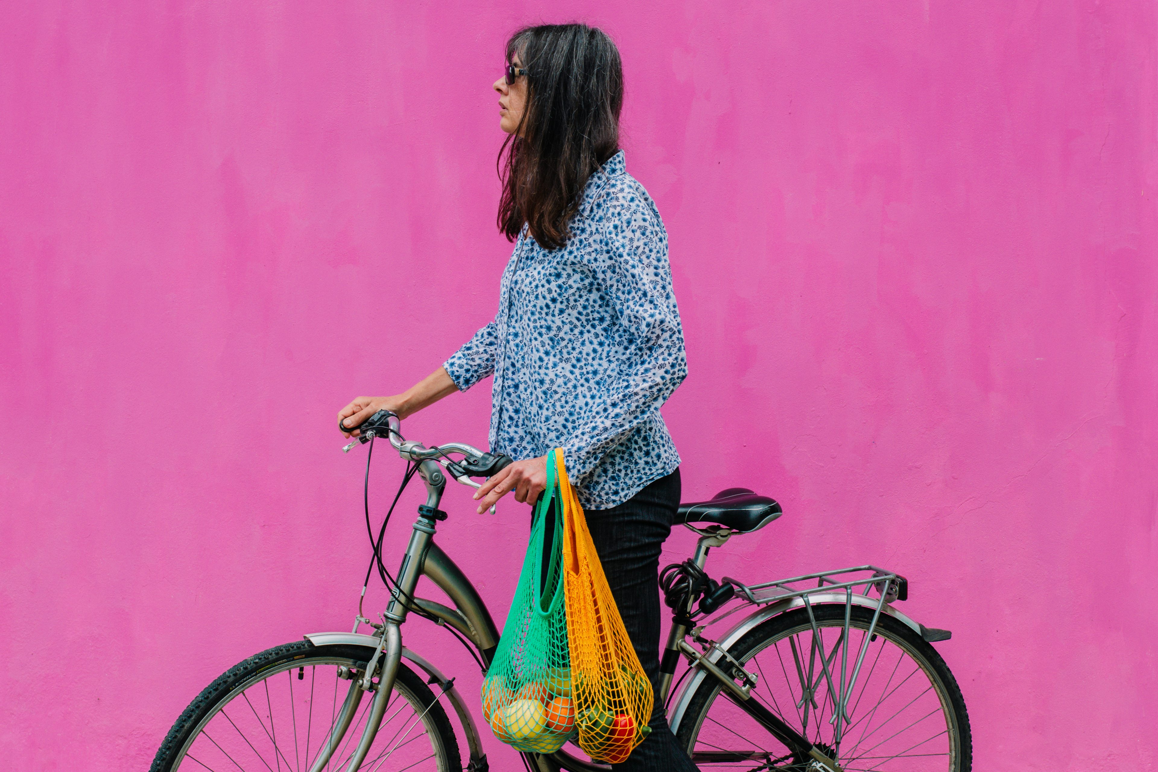 Dark hair woman with bike and net grocery bags with pink wall