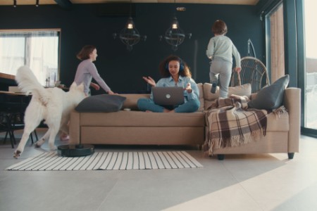 A photograph of a woman working on a sofa as her kids and a dog run around her.