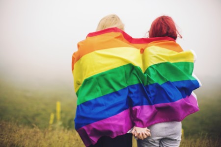 
            Dos mujeres envueltas en la bandera del arco iris cogidas de la mano
        