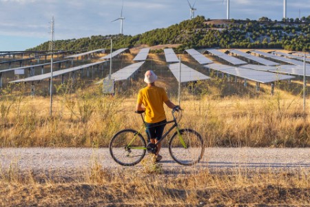 Man from behind leaning on bicycle looking at wind power towers and solar farm in rural setting at sunset