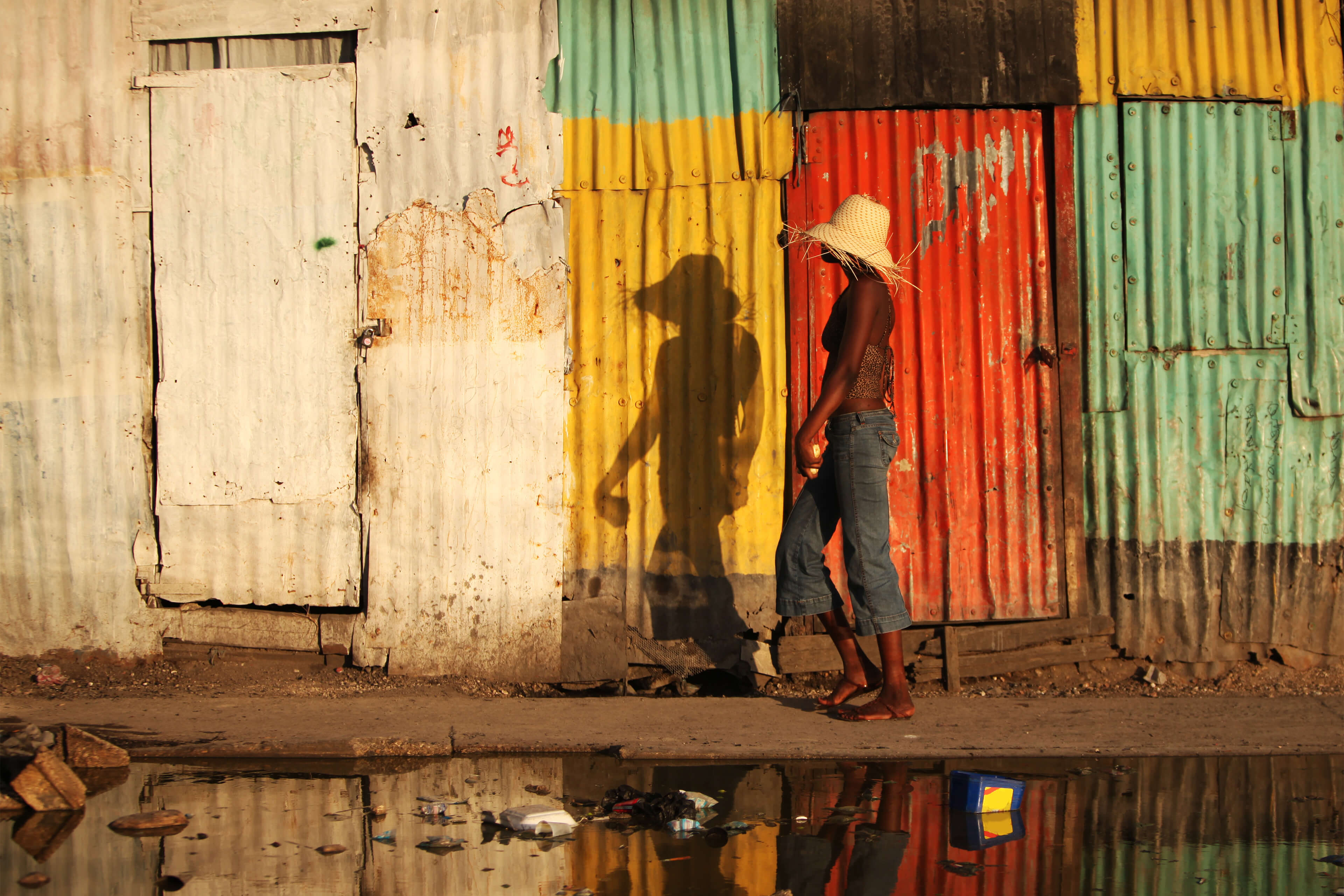 A Haitian woman with a straw hat walks past a rusty corrugated iron wall.