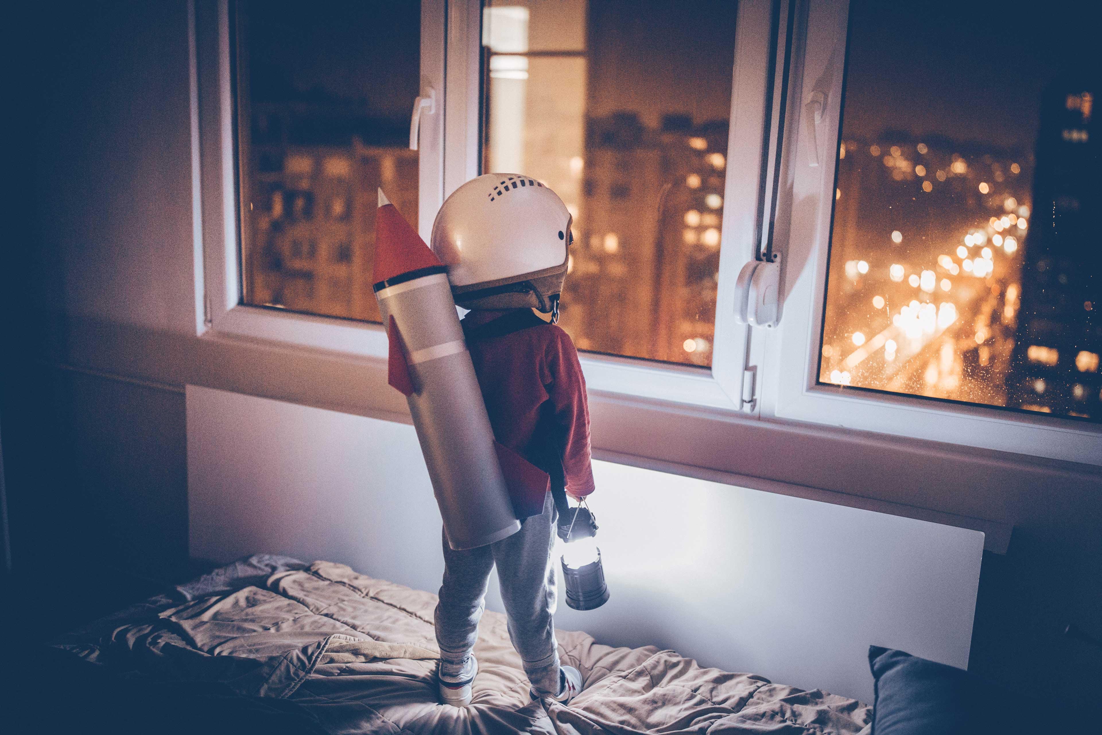 Boy wearing a rocket and standing on the bed and wishing to go to space