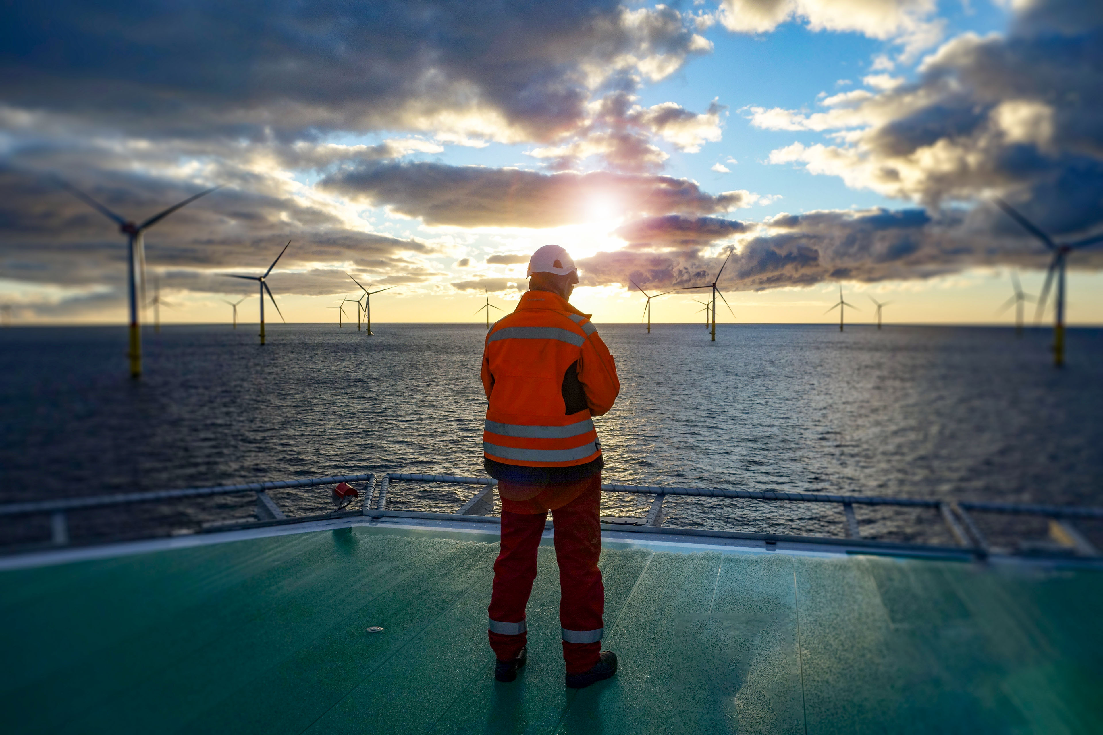 Offshore worker standing on helipad with wind-turbines behind him in sunset