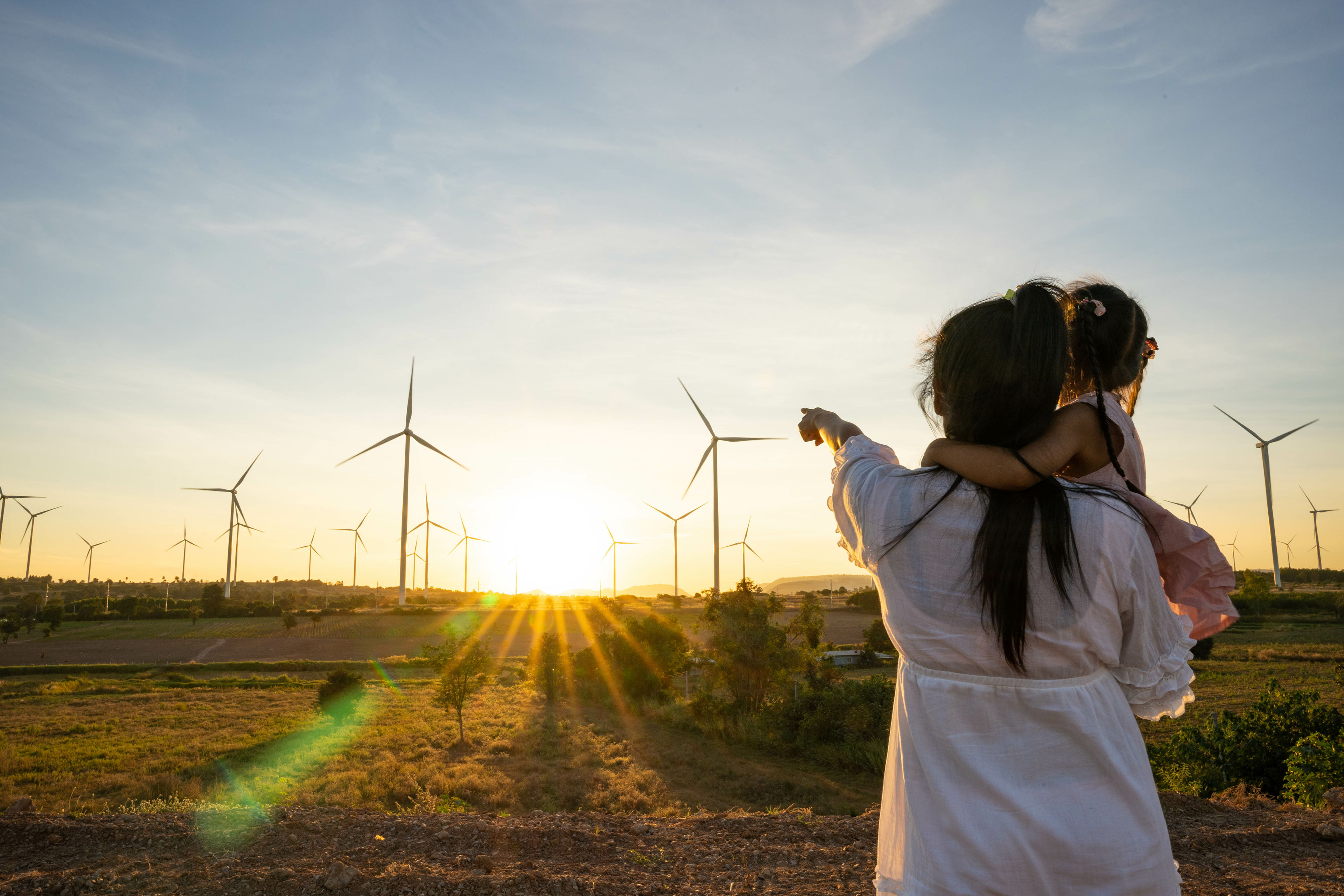 Una mujer cargando a una niña, mientras señala con su mano izquierda unos molinos de energía eólica que se ven en el horizonte