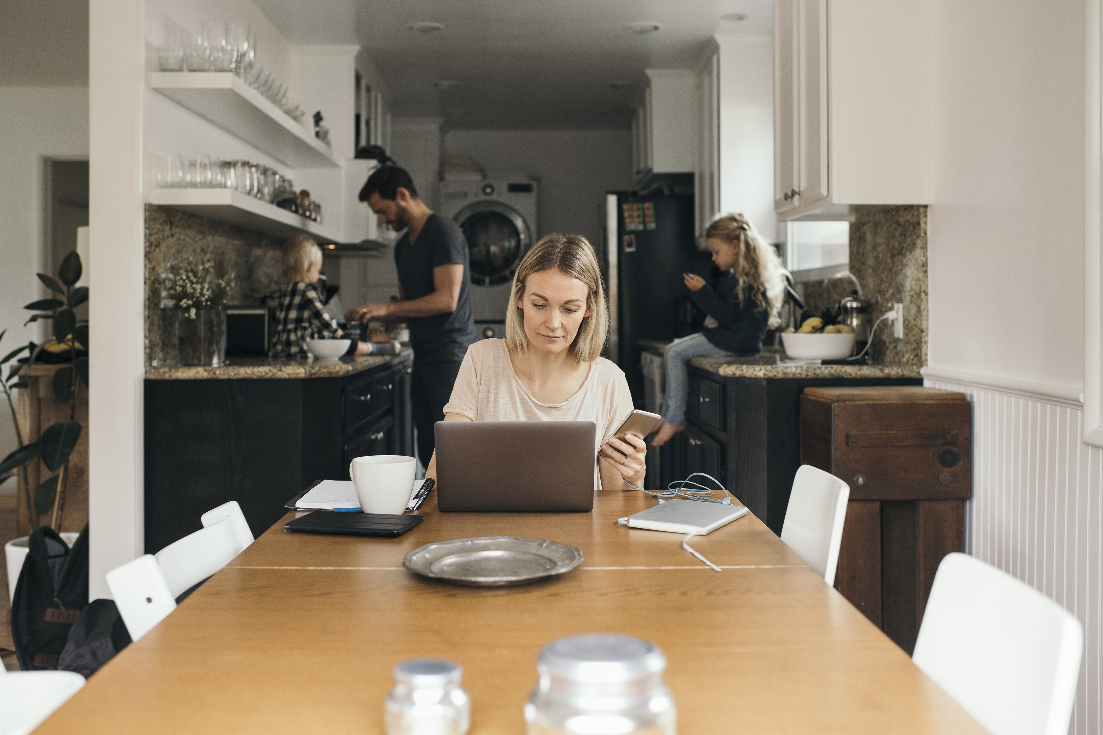 woman holding mobile phone while working on laptop at table with family at home