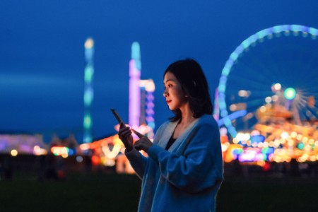 Sonriente joven mirando smartphone en feria de atracciones por la noche