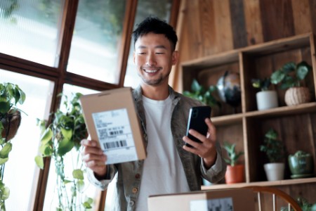Smiling Asian man checking his smartphone as he receives delivered packages from online purchases at home