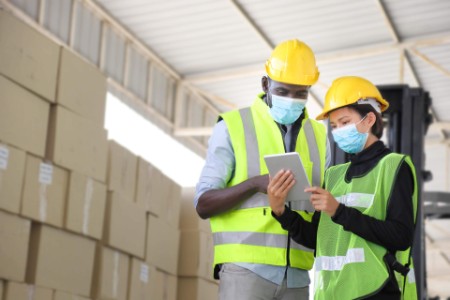 African American and Asian warehouse workers with facial masks using digital tablet