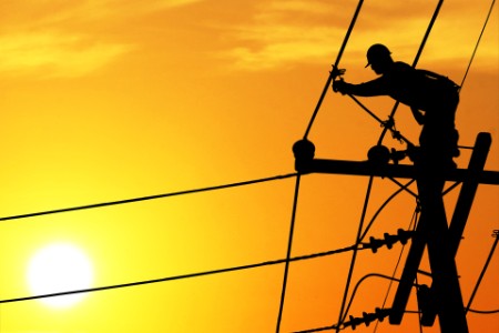 Bild i motljust av en elektriker som arbetar uppe på elstolpar vid solnedgång.