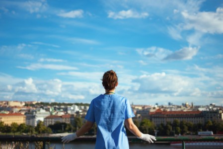 Seen from behind modern physician woman in scrubs with rubber gloves outdoors in the city against sky.