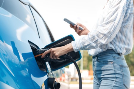 Young adult woman charging electric car, using smartphone