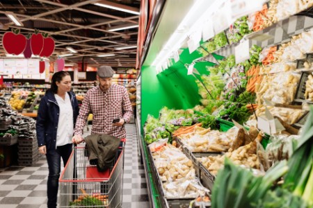 Couple choosing vegetables while using phone in supermarket