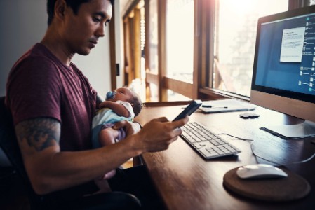 EY - man sitting at desk holding baby and looking at mobile phone