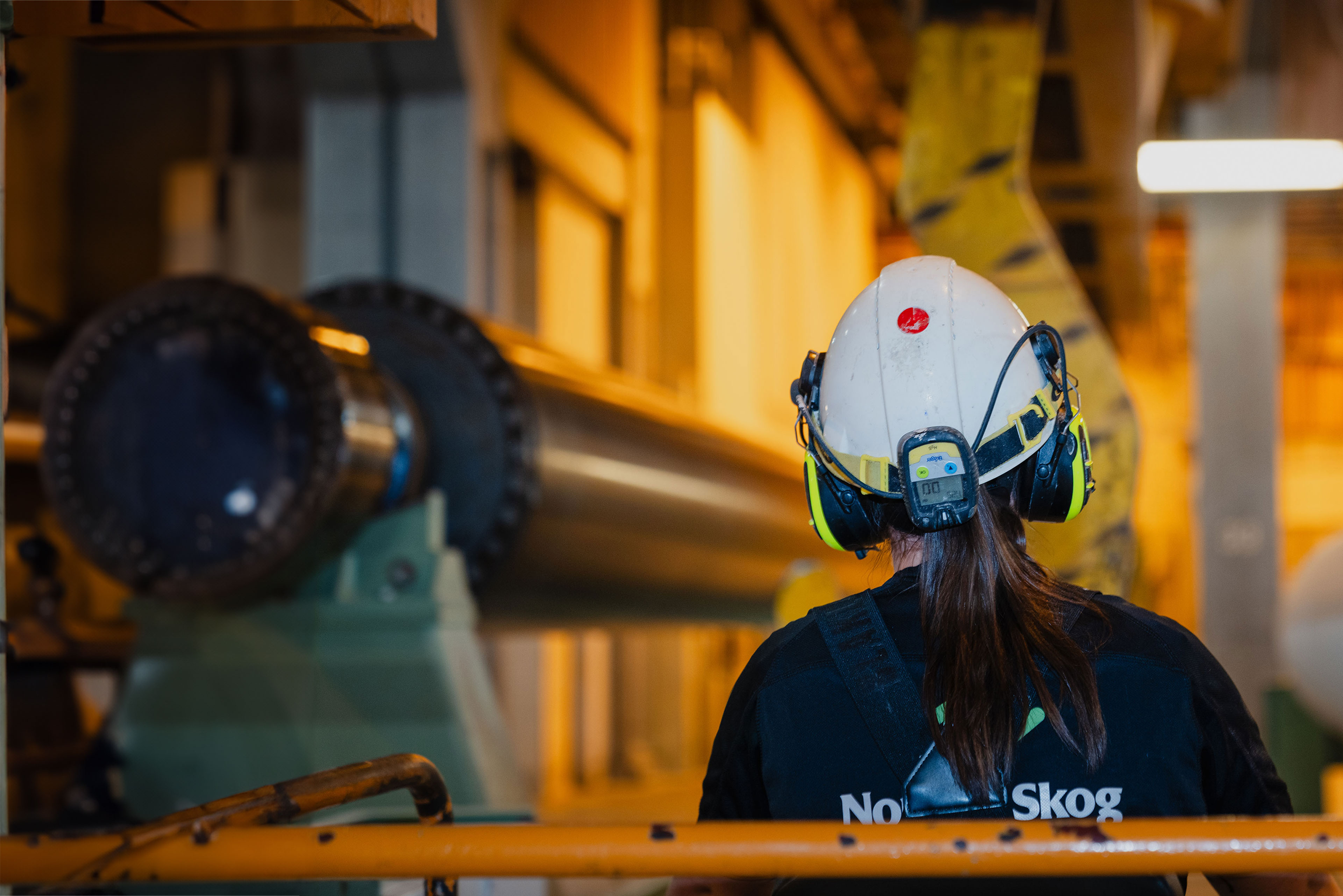 A worker with a helmet, hearing protection and a ponytail stands in front of industrial machinery at Norske Skog.