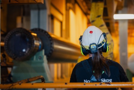 A worker with a helmet, hearing protection and a ponytail stands in front of industrial machinery at Norske Skog.