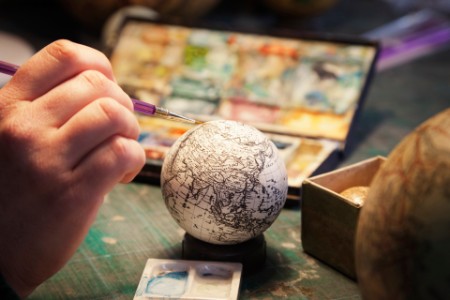 Kartograf maler en liten globus med farger. Hånden holder penselen og legger svarte linjer på en globus på et lyst skrivebord, med vannfargepaletter og en annen globus i bakgrunnen.