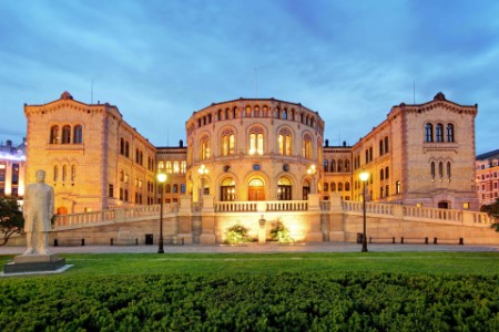 Opplyst Stortinget i Oslo på kveldstid med statue i forgrunnen og frodig grønn plen.