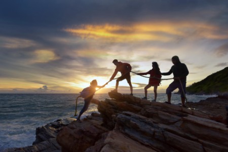 Bild av Mannlige og kvinnelige turgåere klatrer opp silhuettfjellsklippen og en av dem gir hjelpende hånd.
