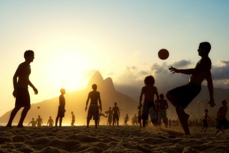 homens jogando futebol na praia e ao fundo o pôr do sol.